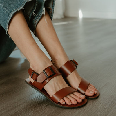 Cognac leather Groundies Kos Sandals with black thermo-rubber soles. One thick strap spans across the base of the toes. Another thick strap goes accross the arch of the foot with a big, black buckle closure. A thinner slingback strap keeps the sandals secure to the foot. Both shoes are shown facing diagonally right on a light skinned woman wearing loose, distressed, light wash jeans sitting on a grey wooden floor. #color_cognac