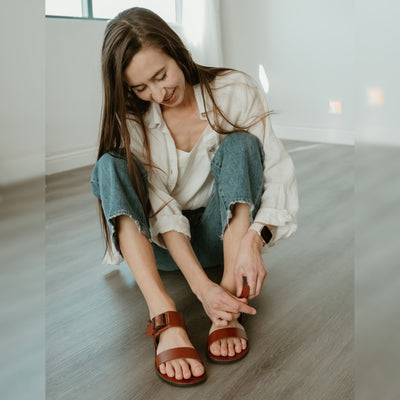 Cognac leather Groundies Kos Sandals with black thermo-rubber soles. One thick strap spans across the base of the toes. Another thick strap goes accross the arch of the foot with a big, black buckle closure. A thinner slingback strap keeps the sandals secure to the foot. Both shoes are shown from the front on a light skinned woman with long brown hair wearing loose, distressed, light wash jeans and an off white button up shirt sitting on a grey wooden floor. #color_cognac