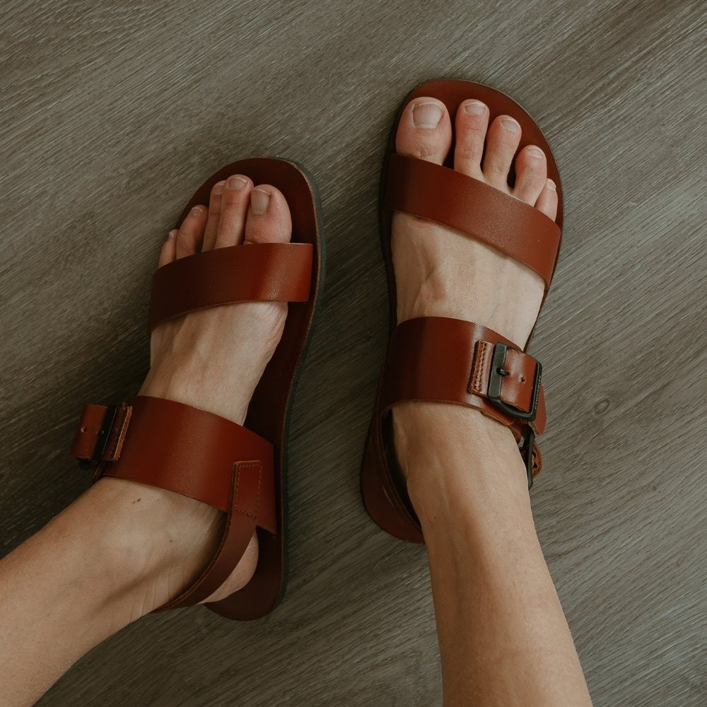 Cognac leather Groundies Kos Sandals with black thermo-rubber soles. One thick strap spans across the base of the toes. Another thick strap goes accross the arch of the foot with a big, black buckle closure. A thinner slingback strap keeps the sandals secure to the foot. Both shoes are shown from above with the left leg tilted to the left on a light skinned woman wearing loose, distressed, light wash jeans sitting on a grey wooden floor. #color_cognac