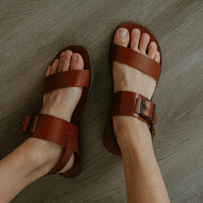 Cognac leather Groundies Kos Sandals with black thermo-rubber soles. One thick strap spans across the base of the toes. Another thick strap goes accross the arch of the foot with a big, black buckle closure. A thinner slingback strap keeps the sandals secure to the foot. Both shoes are shown from above with the left leg tilted to the left on a light skinned woman wearing loose, distressed, light wash jeans sitting on a grey wooden floor. #color_cognac
