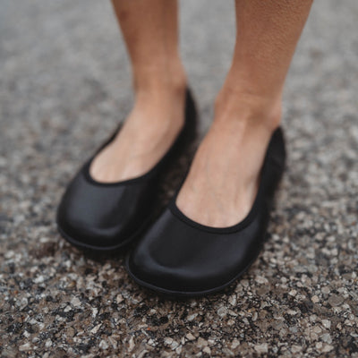 A photo of Groundies Lily Classic flats with a leather upper and black rubber true sense soles. The flats are a smooth leather in a black color with trim around the tops. The interior of the flats is a light beige color. Both flats are shown diagonally from the front left on a womans foot standing on rocky pavement. #color_black