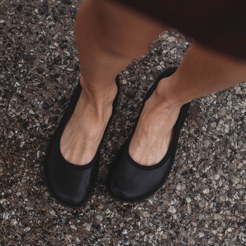 A photo of all black smooth leather Groundies Lily 2 Classic ballet flats. Both flats are shown here from above diagonally left on a womans feet standing on rocky pavement. #color_black