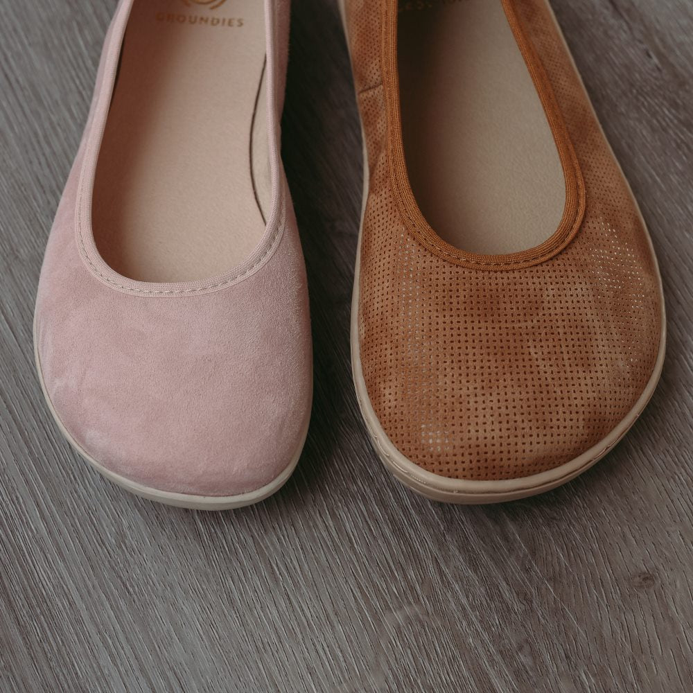 A photo of Groundies Lily Classic flats with a leather upper and tan rubber true sense soles. The flats are a perforated leather in a cognac color with trim around the tops. The interior of the flats is a light beige color. The left flat is shown from the front next to a right light pink Lily Soft flat, on a wood floor. #color_cognac