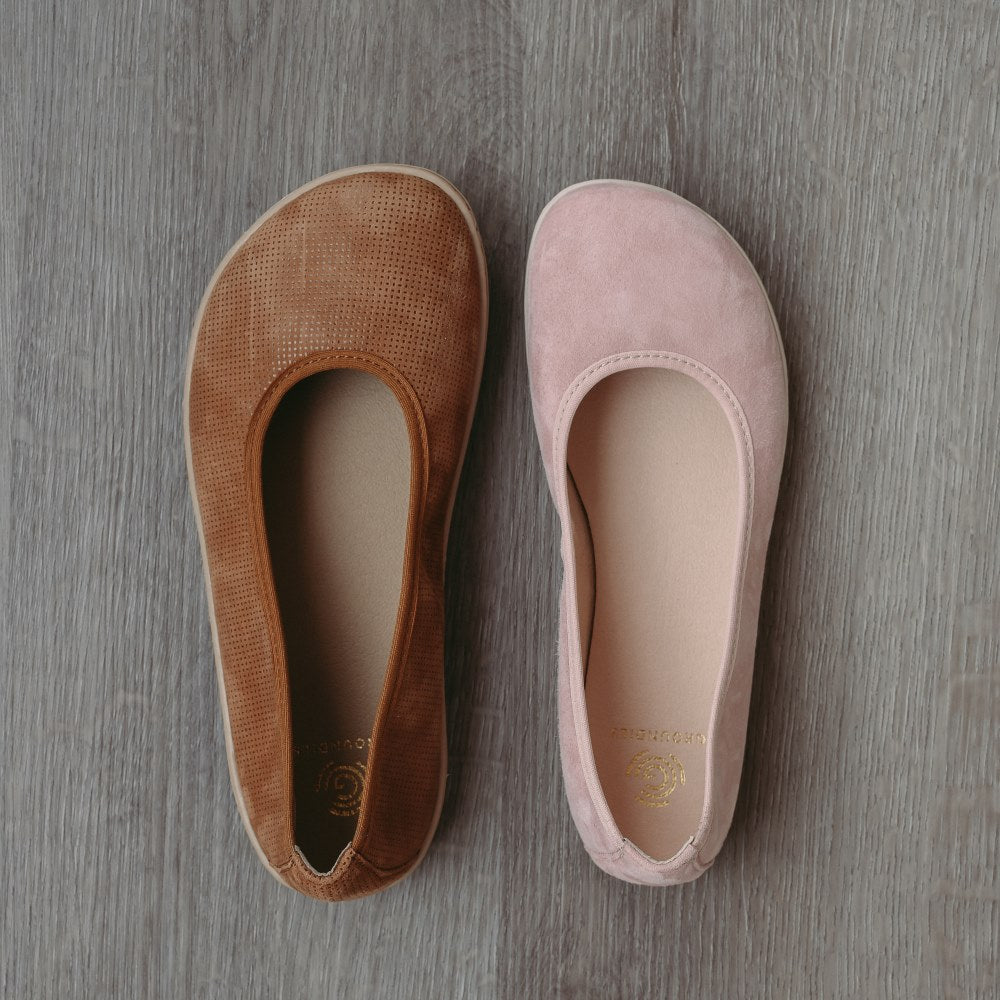 A photo of Groundies Lily Classic flats with a leather upper and tan rubber true sense soles. The flats are a perforated leather in a cognac color with trim around the tops. The interior of the flats is a light beige color. The left flat is shown from above next to a right light pink Lily Soft flat, on a wood floor. #color_cognac