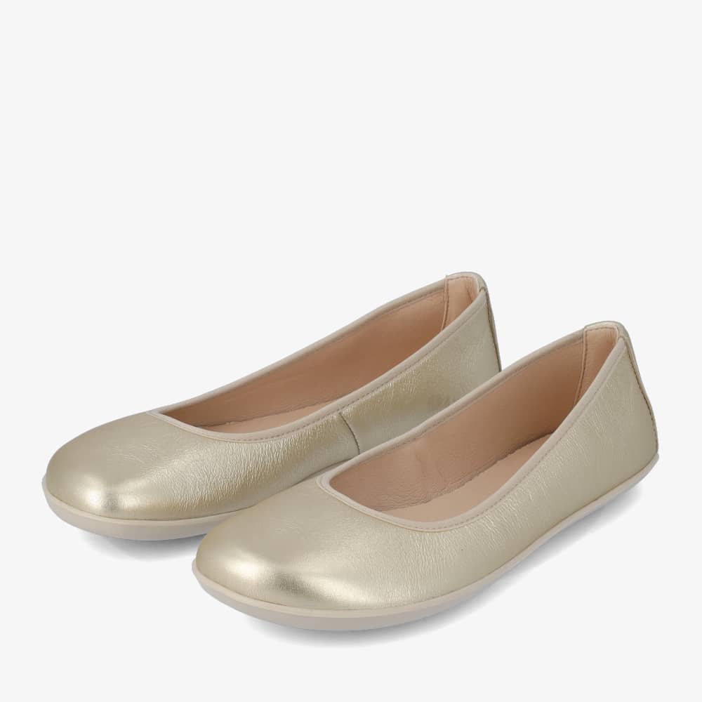 A photo of Groundies Lily Classic flats with a leather upper and white rubber true sense soles. The flats are a perforated leather in a metallic champagne color with trim around the tops. The interior of the flats is a light beige color. Both flats are shown beside each other facing to the left with the left flat on the outside against a white background. #color_metallic-champagne
