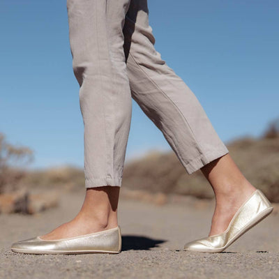 A photo of Groundies Lily Classic flats with a leather upper and white rubber true sense soles. The flats are a perforated leather in a metallic champagne color with trim around the tops. The interior of the flats is a light beige color. Both flats are shown on a woman's feet with a view of her thighs down. The woman is wearing cropped khaki pants and is walking across a desert scene with her right foot in front of her left. #color_metallic-champagne