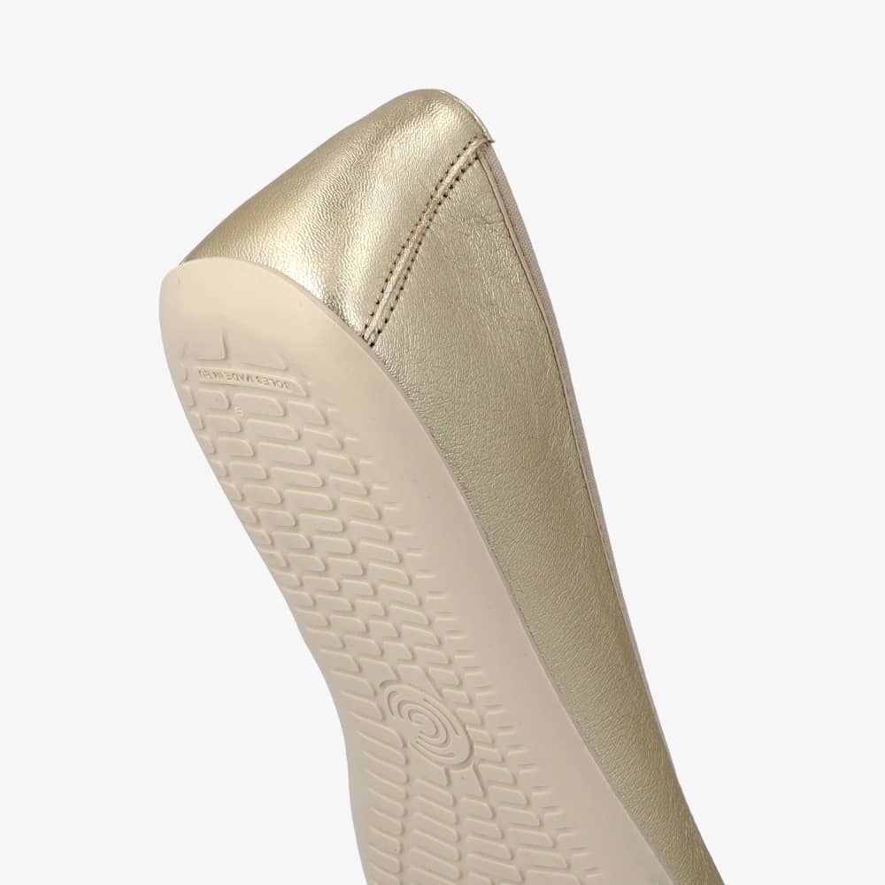 A photo of Groundies Lily Classic flats with a leather upper and white rubber true sense soles. The flats are a perforated leather in a metallic champagne color with trim around the tops. The interior of the flats is a light beige color. The right flat is shown from behind and is tilted downward, displaying the sole and heel of the shoe, on a white background. #color_metallic-champagne