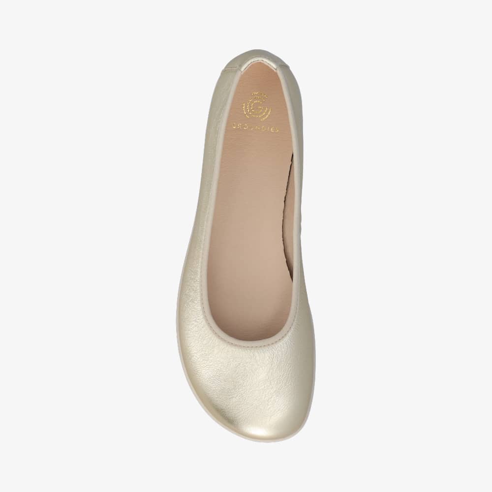 A photo of Groundies Lily Classic flats with a leather upper and white rubber true sense soles. The flats are a smooth leather in a metallic champagne color with trim around the tops. The right flat is shown floating facing downwards against a white background. #color_metallic-champagne