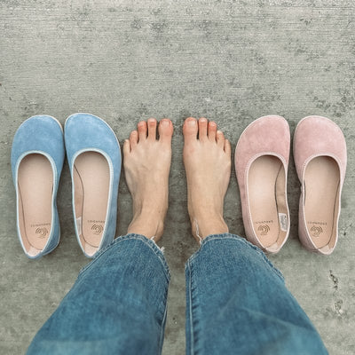 A photo of Groundies Lily Soft flats with a leather upper and cream rubber true sense soles. The flats are a nubuck in a light blue color, the trim around the tops of the flats is a baby blue color and is lighter than the rest of the flats. The interior of the flats is a light beige color. Both the light blue and light pink flats are shown here next to a womans feet standing on cement. #color_light-blue