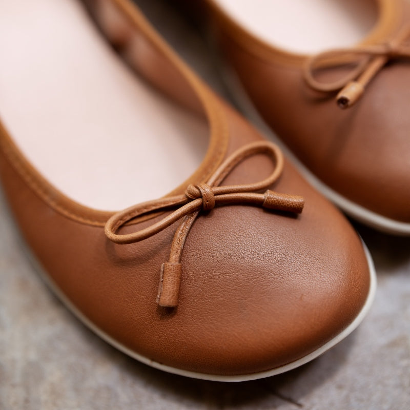 A photo of light brown leather Groundies Magnolia Flats. These classic ballet flats are simple with a leather bow located at the top of the shoe opening. Right shoe is shown close up on the bow detail with the left shoe in the background both on cement. #color_light-brown