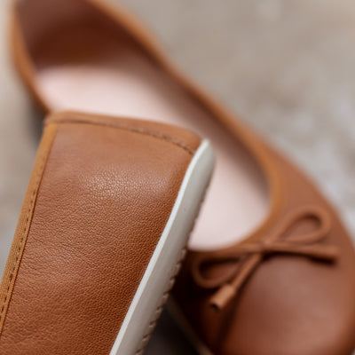 A photo of light brown leather Groundies Magnolia Flats. These classic ballet flats are simple with a leather bow located at the top of the shoe opening. Right shoe is shown close up on the side heel with the left shoe in the background both on cement. #color_light-brown
