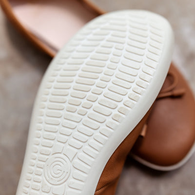 A photo of light brown leather Groundies Magnolia Flats. These classic ballet flats are simple with a leather bow located at the top of the shoe opening. Right shoe is shown close up on the white sole with the left shoe in the background both on cement. #color_light-brown