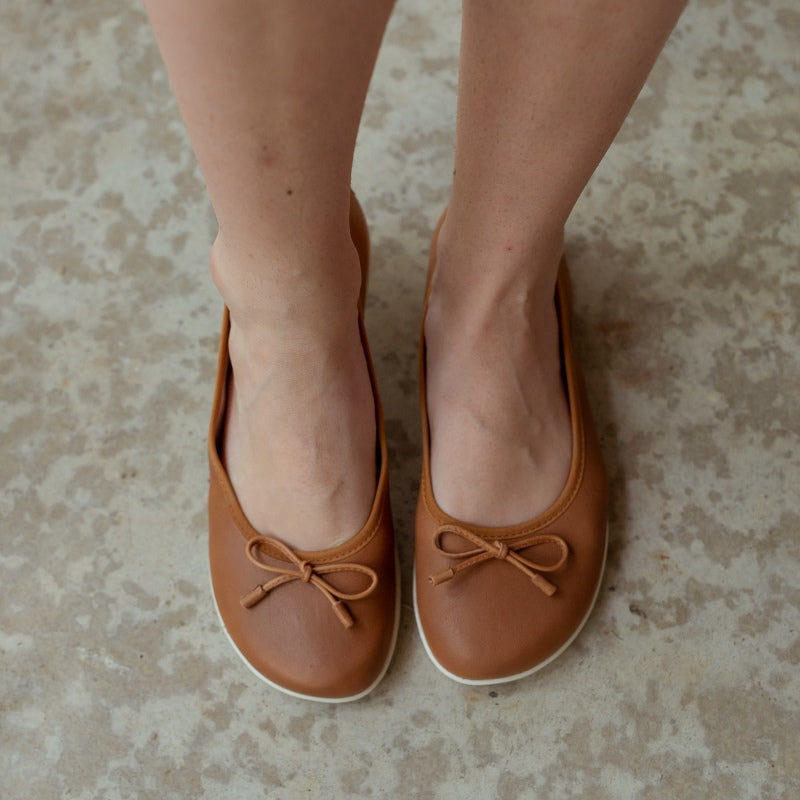 A photo of light brown leather Groundies Magnolia Flats. These classic ballet flats are simple with a leather bow located at the top of the shoe opening. Shoes are shown from the top down on a womans feet standing on cement. #color_light-brown