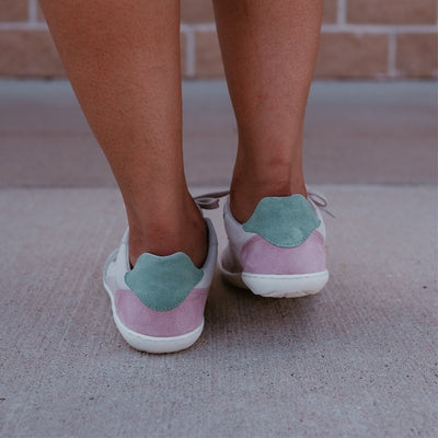 Beige Pink Groundies Nova GO1 leather Sneakers. The sneakers are an off white base leather with suede beige, green, and pink color blocks. Both shoes are shown from behind on a dark-skinned woman standing on pavement with a brick wall behind. #color_beige-pink
