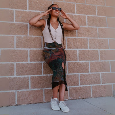 Beige Pink Groundies Nova GO1 leather Sneakers. The sneakers are an off white base leather with suede beige, green, and pink color blocks. Both shoes are shown facing right on a dark-skinned woman standing on pavement leaning on a brick. She is wearing a deep-colored, form-fitting skirt with a side slit tucked into a beige vest and dark, cat-eye sunglasses. #color_beige-pink