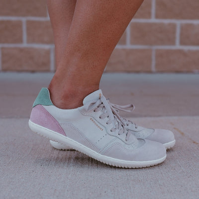 Beige Pink Groundies Nova GO1 leather Sneakers. The sneakers are an off white base leather with suede beige, green, and pink color blocks. Both shoes are shown facing right on a dark-skinned woman standing on pavement with a brick wall behind. #color_beige-pink