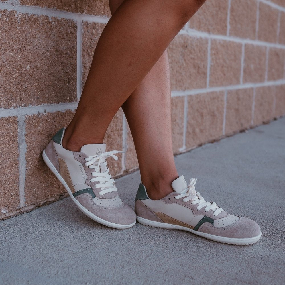 Groundies Nova GO1 leather Sneakers. Sneakers are a cream leather base with suede beige, sage green, and tan color blocks. Both shoes are shown facing diagonally right on a dark-skinned woman standing on pavement and leaning against a brick wall. #color_beige-sage-green