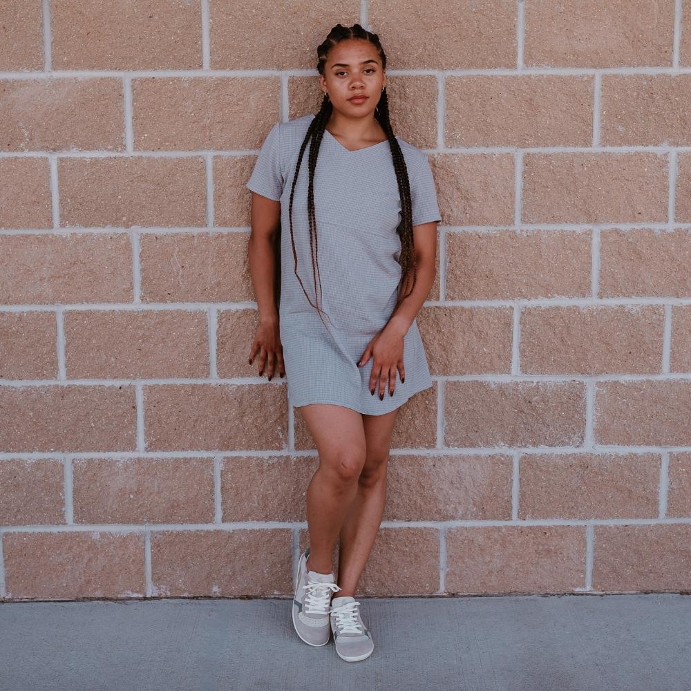 Groundies Nova GO1 leather Sneakers. Sneakers are a cream leather base with suede beige, sage green, and tan color blocks. Both shoes are shown from the front on a dark-skinned woman with long, black braids wearing a short, grey t-shirt dress standing on pavement and leaning against a brick wall. #color_beige-sage-green