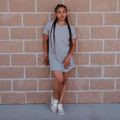 Groundies Nova GO1 leather Sneakers. Sneakers are a cream leather base with suede beige, sage green, and tan color blocks. Both shoes are shown from the front on a dark-skinned woman with long, black braids wearing a short, grey t-shirt dress standing on pavement and leaning against a brick wall. #color_beige-sage-green