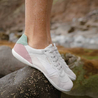 A photo of Groundies Nova GO1 Sneakers made with soft leather and white rubber soles. The sneakers have Beige, Green, and Pink color blocks. Both shoes are shown from the right side on a woman's feet with a view of her shins down. The woman is standing on a large rock demonstrating the flexibility of the soles. #color_beige-green-pink
