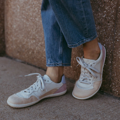 A photo of Groundies Nova GO1 Sneakers made with soft leather and white rubber soles. The sneakers have Beige, Periwinkle, and Pink color blocks. Both shoes are shown here on womens feet from the left diagonal with woman wearing wide legged blue jeans. Left foot is propped up against a brown cement wall while the right foot rests on the ground. #color_beige-lavender-pink