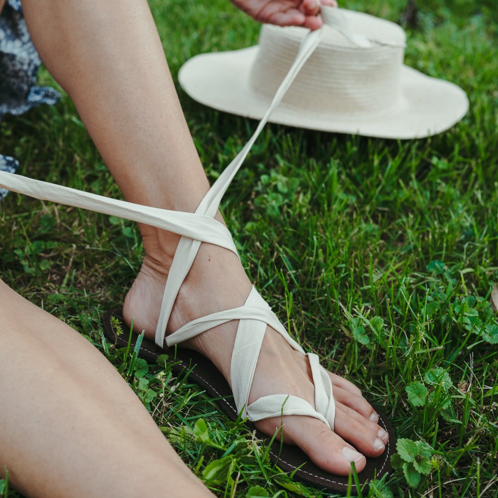 A photo of Juuri ribbon sandals made from leather, viscose, and rubber soles. The sandals have a brown footbed and long sand laces. Left sandal is shown being tied from the left side on a tan woman sitting in grass with a tan wide-brim hat on the grass behind. #color_brown-sand