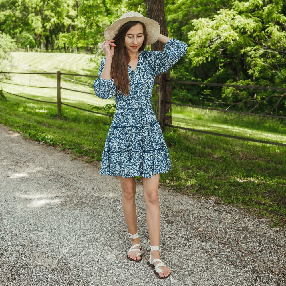 A photo of Juuri ribbon sandals made from leather, viscose, and rubber soles. The sandals have a brown footbed and long sand laces. Shoes are shown on a tan woman with long brown hair wearing a mid-thigh blue and white floral cottage-style dress with 3 quarter-lenth sleeves. She is wearing a tan wide-brim hat, standing on a grey paved road with a wooden fence, grass, and woods in the background. #color_brown-sand