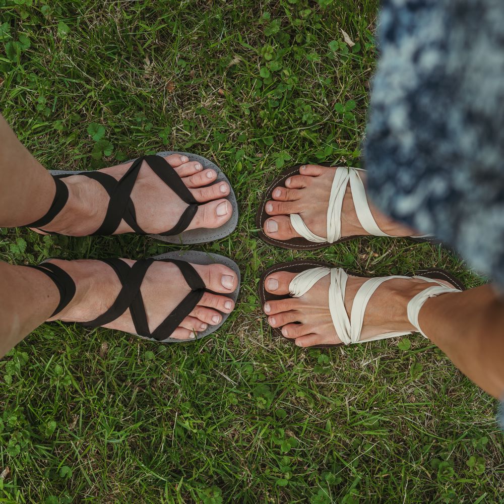 A photo of Juuri ribbon sandals made from leather, viscose, and rubber soles. The sandals have a grey footbed and long black laces. Sandals are shown from above on a womans feet standing in grass with another woman wearing a brown/beige colorway of the sandal standing opposite her. #color_grey-black