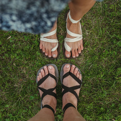 A photo of Juuri ribbon sandals made from leather, viscose, and rubber soles. The sandals have a grey footbed and long black laces. Sandals are shown from above on a womans feet standing in grass with another woman wearing a brown/beige colorway of the sandal standing opposite her. #color_grey-black