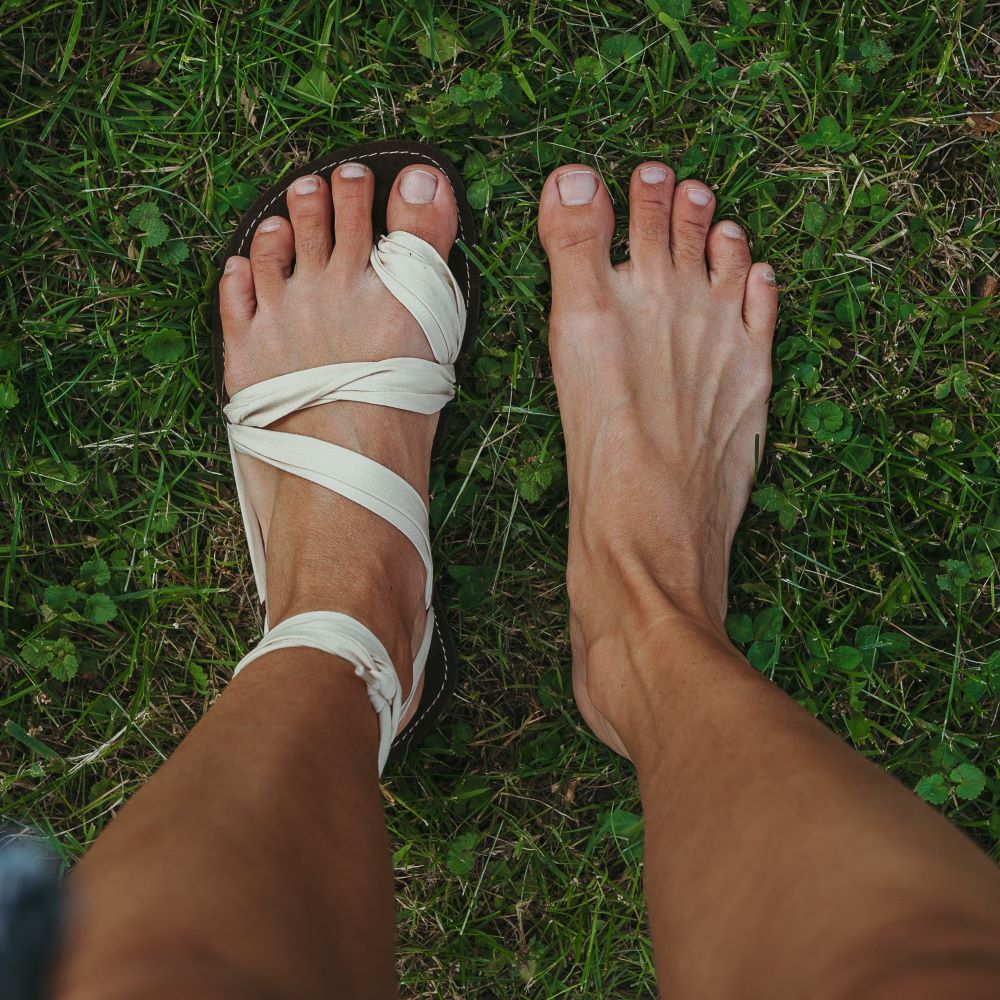 A photo of Juuri ribbon sandals made from leather, viscose, and rubber soles. The sandals have a brown footbed and long sand laces. Left sandal is shown from above next to a right bare foot on a tan woman standing in grass. #color_brown-sand