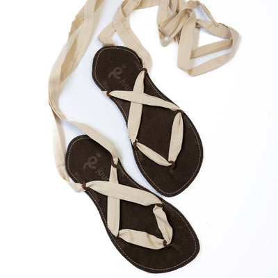 A photo of Juuri ribbon sandals made from leather, viscose, and rubber soles. The sandals have a brown footbed and long sand laces. Both sandals are shown from above facing diagonally right against a white background. #color_brown-sand