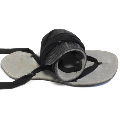 A photo of Juuri ribbon sandals made from leather, viscose, and rubber soles. The sandals have a grey footbed and long black laces. Both sandals are shown, the left sandal is shown on top of the right rolled into a ball to shown flexibility against a white background. #color_grey-black