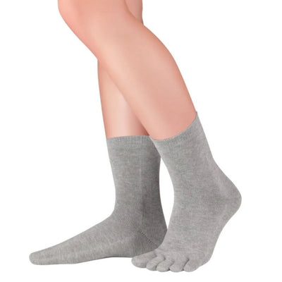 A photo of Knitido essential midi crew toe socks made with cotton, nylon, and elastane. The socks are a grey color. A woman is shown from the knee down wearing the midi crew socks, she is facing to the right with the heel of her left foot lifted off the floor against a white background. #color_grey