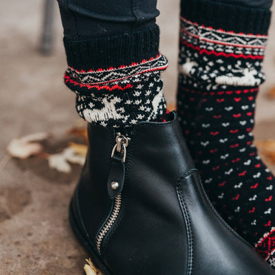 A photo of the Knitido Hossa reindeer sweater toe socks, which are cotton and wool blend. The socks are black in color with beige and red festive details all over the sock and reindeer around the ankle. The toes and heels are beige. Both socks are shown on a woman's feet from the right side with a view of her shins down.The woman is wearing black leggings tucked into the socks and a black boot on her right foot, and she is standing on a cement floor with wet leaves on it. #color_black-beige