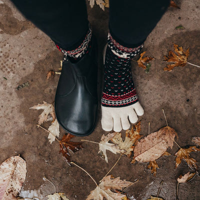 A photo of the Knitido Hossa reindeer sweater toe socks, which are cotton and wool blend. The socks are black in color with beige and red festive details all over the sock and reindeer around the ankle. The toes and heels are beige. Both socks are shown on a woman's feet from above with a view of her knees down.The woman is wearing black leggings tucked into the socks and a black boot on her right foot, and she is standing on a cement floor with wet leaves on it.  #color_black-beige