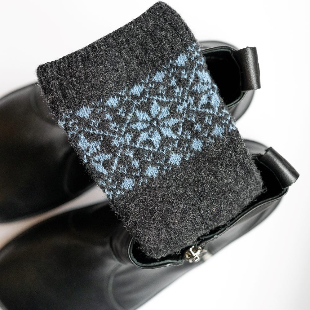 A photo of anthracite and blue knitted toe socks, they have blue detailed snowflake design around the mid-calf. One of the socks is shown folded up over a pair of black chelesa boots with a gold zipper against a white background. #color_anthracite-light-blue