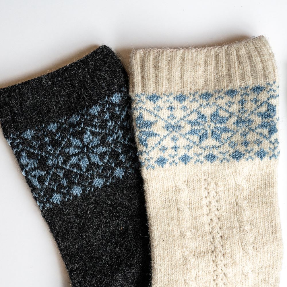 A photo of beige and blue knitted toe socks, they have blue detailed snowflake design around the mid-calf. One sock in the beige/blue color is shown and one sock in the anthracite/blue color is laying stacked with it up close against a white background. #color_beige-blue