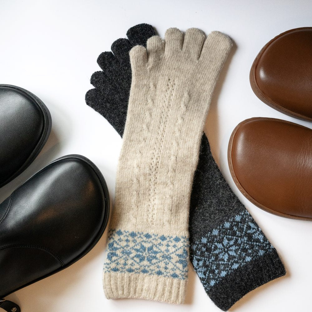 A photo of beige and blue knitted toe socks, they have blue detailed snowflake design around the mid-calf. One sock in the beige/blue color is shown and one sock in the anthracite/blue color is laying stacked with it against a white background with the toe box of a brown and black shown in each side. #color_beige-blue