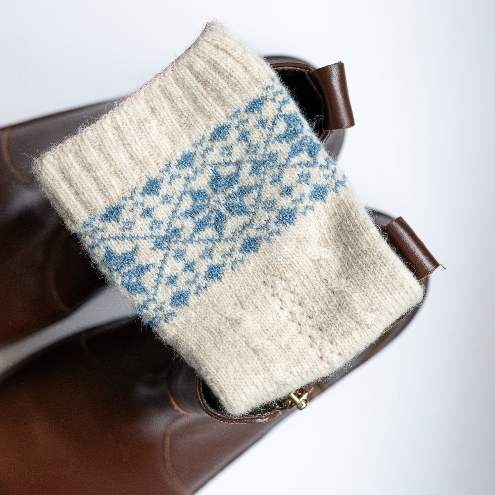 A photo of beige and blue knitted toe socks, they have blue detailed snowflake design around the mid-calf. One of the socks is shown folded up over a pair of brown chelesa boots with a gold zipper against a white background. #color_beige-blue