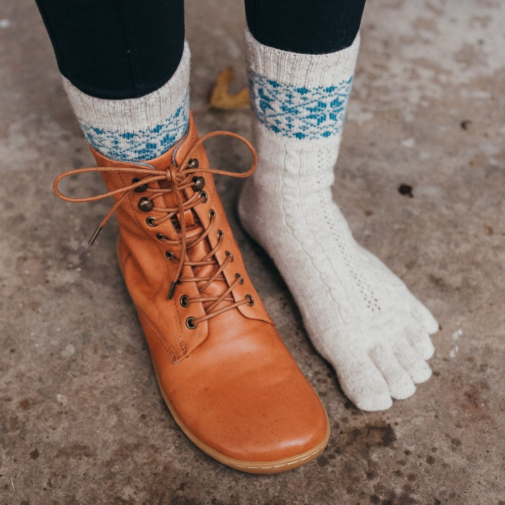 A photo of beige and blue knitted toe socks, they have blue detailed snowflake design around the mid-calf. A woman is shown standing to the right side on concrete wearing black leggings, one carmel boot, and the beige/blue knitido socks. #color_beige-blue