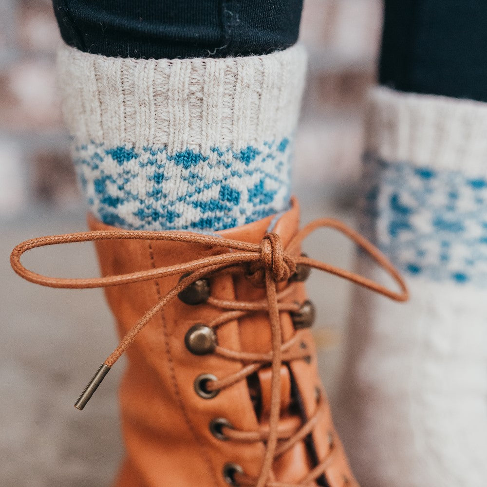 A photo of beige and blue knitted toe socks, they have blue detailed snowflake design around the mid-calf. An up close photo is shown of a women’s right leg wearing the beige/blue snowflake socks, black leggings, and the carmel boots. #color_beige-blue