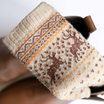 A photo of the Knitido Hossa reindeer sweater toe socks, which are cotton and wool blend. The socks are beige in color with brown and yellow festive details all over the sock and reindeer around the ankle. The toes and heels are brown. One of the socks is shown folded displaying the reindeer detail over a pair of brown boots on a white background.  #color_beige-brown