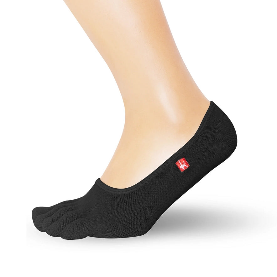 A photo of Knitido track and trail footies socks made with cotton, nylon, polyester, and elastane. The socks are a black color. One sock is shown on a foot from mid leg down facing to the right side with it’s heel lifted slightly off the floor against a white background. #color_black