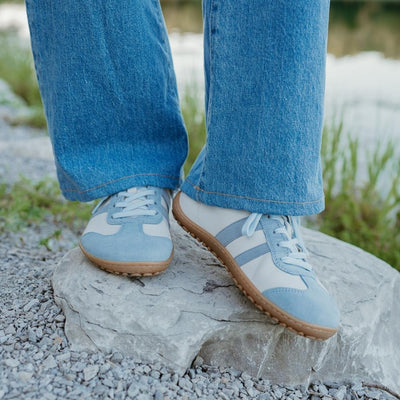 Off White Koel Ila sneakers with two diagonal, baby blue stripes on either side and a baby blue color block at the top of the heel with natural rubber colored soles. A light blue leather T-shape design is present at the top of the toe box with the same color edging the light blue laces. Left shoe is shown from the front while right shoe is angled to the right on a woman wearing long, light-wash, wide-leg jeans standing on a rock by the water and grass. #color_aqua