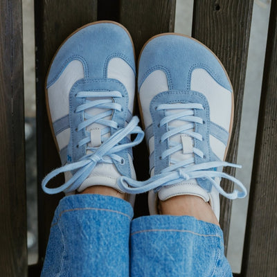 Off White Koel Ila sneakers with two diagonal, baby blue stripes on either side and a baby blue color block at the top of the heel with natural rubber colored soles. A light blue leather T-shape design is present at the top of the toe box with the same color edging the light blue laces. Both shoes are shown from above on a woman wearing long, light-wash, wide-leg jeans sitting on a dark-brown bench. #color_aqua