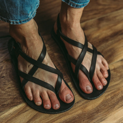 A black leather strappy sandal shown on a womans feet from the front right diagonal against a tan and dark brown wood floor. #color_black