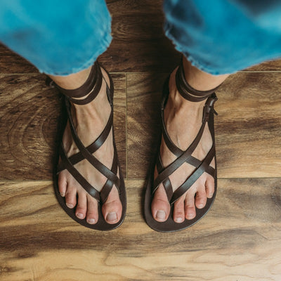 a dark brown leather strappy sandal shown on a womans feet from above against a tan and dark brown wood floor #color_brown