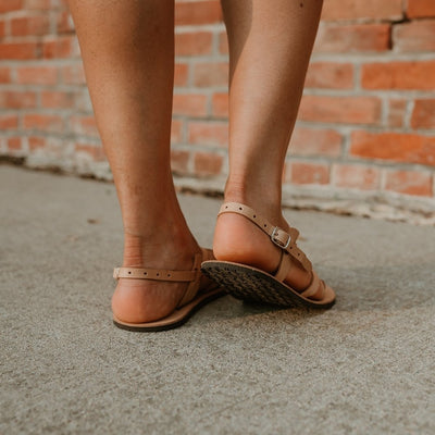 Tan, leather, strappy sandal with a silver buckle in the back. Both sandals are shown from behind on a tan woman standing on a cement path next to a brick building. #color_tan