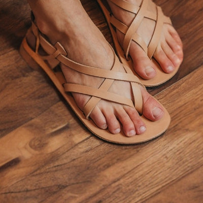 Tan, leather, strappy sandal with a silver buckle in the back. Both sandals are shown facing diagonally right on a light-skinned woman sitting on a brown wooden floor. #color_tan