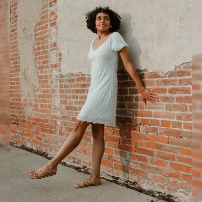 Tan, leather, strappy sandal with a silver buckle in the back. Both sandals are shown on a tan woman with short, black, curly hair wearing a short, white, whimsical dress leaning back against a brick wall. #color_tan
